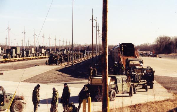 Army vehicles are loaded onto rail transport cars in the foreground. Meanwhile, in the background, army vehicles sit on flat cars ready to roll out.